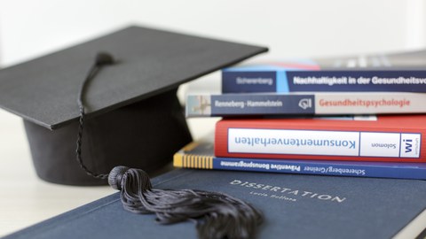 Man sieht eine Dissertation, einen Stapel Bücher und einen Doktorhut